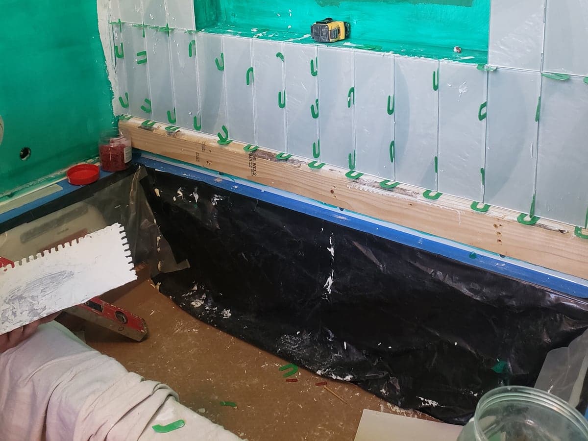 Bathroom shower wall under construction with waterproofing membrane, backer board, tile spacers, and a tub surround.